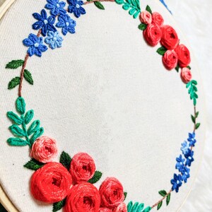 May include: A white embroidery hoop with a floral wreath design. The wreath is made of red, pink, and blue flowers and green leaves.