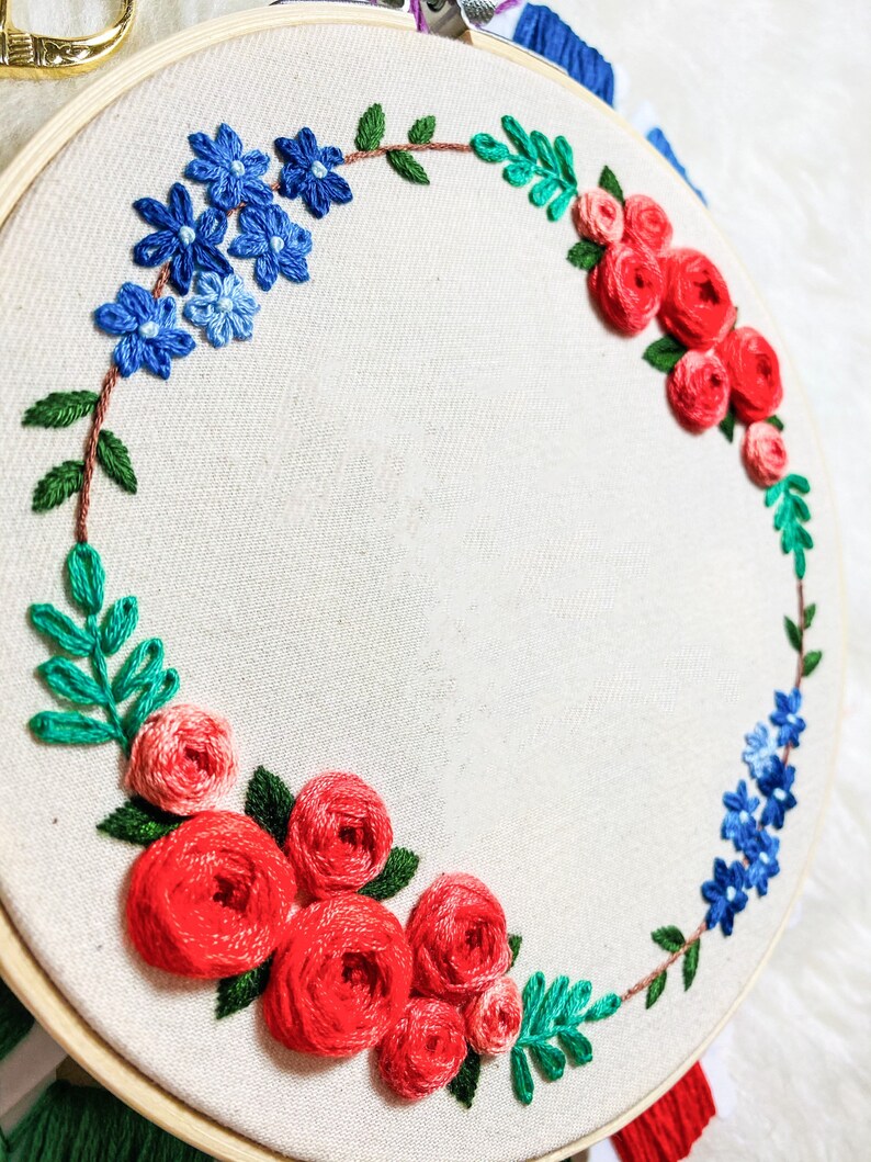 May include: A white embroidery hoop with a floral wreath design. The wreath is made of red, pink, and blue flowers and green leaves.