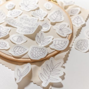 Pode incluir: Uma cole&ccedil;&atilde;o de patches de tecido branco com desenhos de folhas bordadas, incluindo bolotas, pinhas e o texto "Fall Breeze Autumn Leaves". Os patches est&atilde;o dispostos sobre uma superf&iacute;cie de tecido bege e um aro de bordado de madeira.