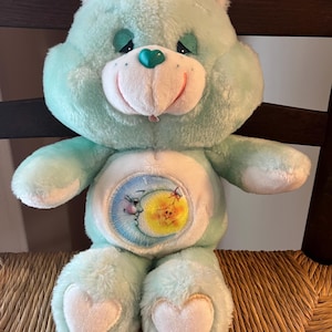 May include: A light blue plush bear with a white belly featuring a moon and sun design. The bear has a green heart-shaped nose, a smiling mouth, and white heart-shaped accents on its feet. It has a green halo and is sitting on a woven chair.