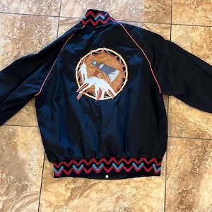 May include: Black satin jacket with a Native American-inspired design. The back features a circular emblem with a dreamcatcher and feather motif. Red and blue zigzag patterns adorn the cuffs and hem. The jacket has a stand-up collar and a button closure.