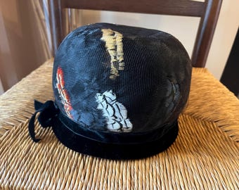 May include: A black cloche hat with a velvet band and a decorative bow. The hat features embroidered details in red, white, and gold. The hat is sitting on a woven surface.