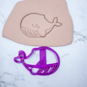 May include: A purple plastic whale cookie cutter. The cutter is shown on a white marble surface with a light brown impression of the whale in modeling clay.
