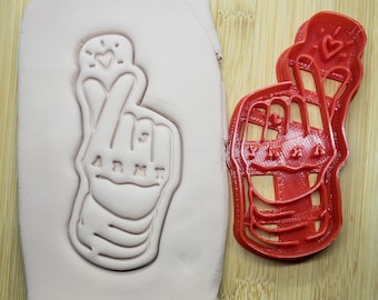 May include: Red plastic cookie cutter shaped like a hand making the Korean heart sign with the word "ARMY" written on the palm. The cutter is next to a pink clay impression of the cutter.
