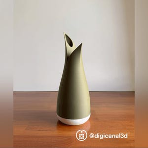 May include: A modern, olive green vase with a white base. The vase has a unique, tulip-like shape with two upward-curving sections at the top. The vase is displayed on a wooden surface.