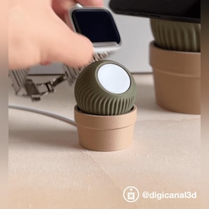 Puede incluir: Un cargador verde para Apple Watch en un pequeño soporte con forma de maceta de color beige. El cargador tiene una superficie de carga blanca y un diseño acanalado. Un Apple Watch se sostiene sobre el cargador. El cargador está conectado a un cable blanco.