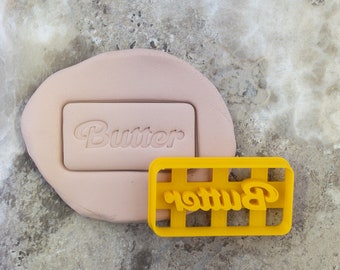 May include: Yellow plastic cookie cutter with the word "Butter" in a stylized font. The cutter has a grid pattern.