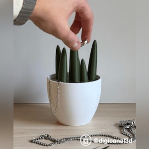 Puede incluir: Un joyero de cerámica blanca con puntas verdes, sosteniendo un anillo plateado. Una cadena de plata y un colgante plateado están sobre la superficie de madera. El joyero está diseñado para organizar y exhibir anillos y collares.