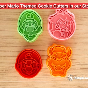 Puede incluir: Cuatro cortadores de galletas de pl&aacute;stico con forma de personajes de la franquicia de videojuegos Super Mario. Los cortadores son verdes, rosas, rojos y naranjas. Los personajes son Luigi, la princesa Peach, Mario y Bowser.