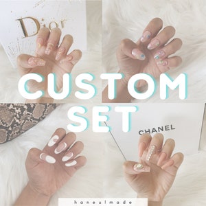 以下が含まれることがあります： さまざまなデザインの、カスタムメイドのつけ爪が4セットあります。爪は形とサイズが異なり、金色の星、月、ハート、ラインストーンで飾られています。画像の中央には、大きな白い文字で「CUSTOM SET」と書かれています。