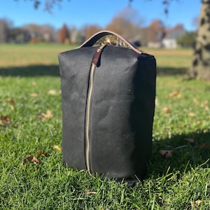 May include: A black canvas shoe bag with a zipper closure and a brown leather handle. The bag is sitting on a green grassy surface.
