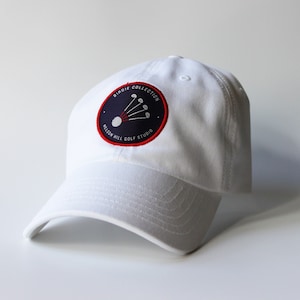 Pode incluir: Boné de beisebol branco com um remendo bordado em azul marinho e vermelho que diz "Birdie Collection Nelson Hill Golf Studio" e apresenta uma bola de golfe com cinco tacos de golfe.