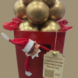 Könnte beinhalten: Eine rote Geschenkbox mit goldenen Kugeln und rotem Seidenpapier. Eine Elfenpuppe schaut aus der Box. Ein Holzanhänger mit der Aufschrift "EXPRESS ELF DELIVERY A Special Message From Santa Claus. Just For You."