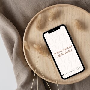 以下が含まれることがあります： 黒い画面に「Support your local caffeine dealers」というテキストが表示された白いスマートフォン。周囲には茶色のフォントで「COFFEE」という単語が並んでいます。スマートフォンは、乾燥した草が敷かれた木製のトレイの上に置かれています。