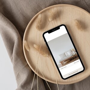 May include: A black smartphone displaying a website on a wooden tray with dried grass. The website features a photo of a white sofa with pillows and a woven basket. The text on the website reads "Lorem Ipsum" and "Shop all goods".