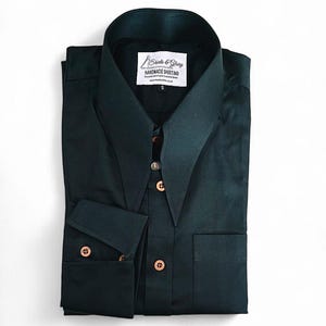 Puede incluir: Una camisa de manga larga de color verde oscuro con cuello puntiagudo y un solo bolsillo en el pecho. La camisa tiene una etiqueta que dice "Shirts & Glory Handmade Shirting".