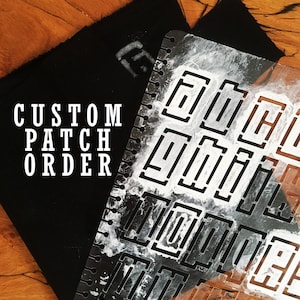 Pode incluir: Um remendo de tecido preto com o texto "CUSTOM PATCH ORDER" impresso em branco. Um estêncil de plástico transparente com um padrão geométrico é colocado sobre o tecido.