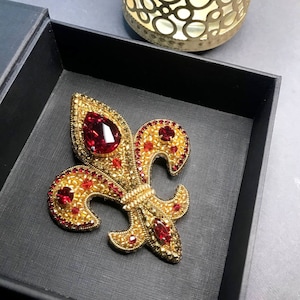 May include: A gold and red jeweled fleur-de-lis brooch in a black velvet box.