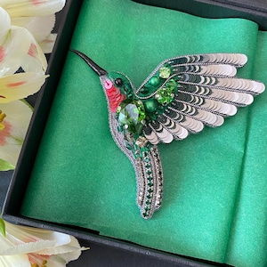 May include: A handcrafted hummingbird brooch with a detailed design. The bird features green, red, and silver accents, with a black beak and sparkling embellishments. Presented in a black gift box with green tissue paper, alongside white flowers.