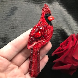 Op de afbeelding: Een rode kardinaal broche gemaakt van pailletten en kralen. De kardinaal heeft een zwarte snavel en een rood lichaam met een grote rode edelsteen in het midden van zijn borst.