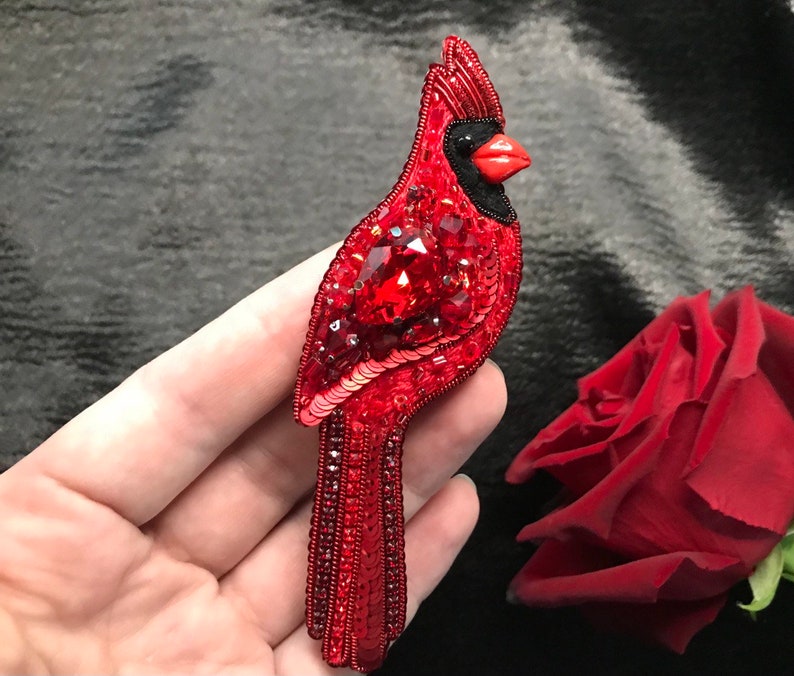 Op de afbeelding: Een rode kardinaal broche gemaakt van pailletten en kralen. De kardinaal heeft een zwarte snavel en een rood lichaam met een grote rode edelsteen in het midden van zijn borst.