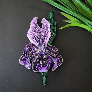 May include: A purple and green beaded iris flower brooch. The flower is made of sequins, beads, and crystals. The stem is green and has leaves.