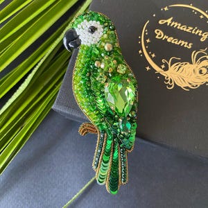 Pode incluir: Broche de papagaio verde, intrincadamente adornado com contas e lantejoulas verdes de várias tonalidades. Apresenta uma grande gema verde facetada e um bico preto. O broche está sobre um fundo escuro, com folhas verdes ao fundo.
