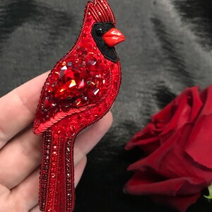 Op de afbeelding: Een rode kardinaalvogel broche gemaakt van pailletten en kralen. De vogel heeft een zwarte kop en een rode snavel.