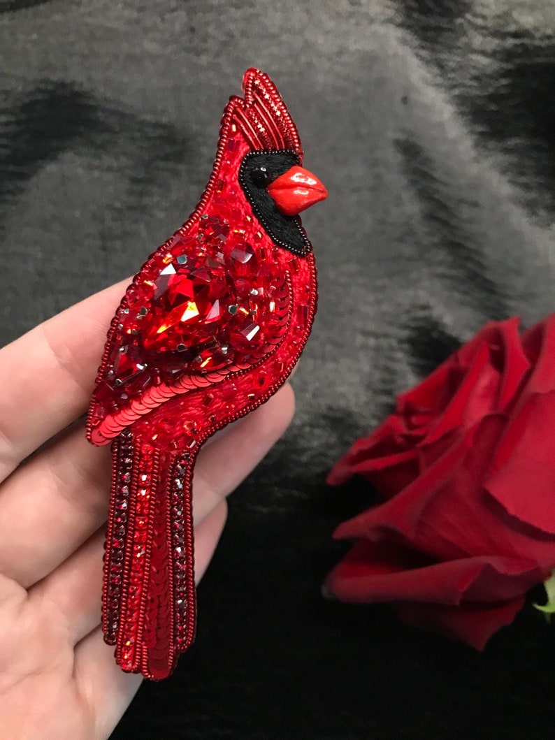 Op de afbeelding: Een rode kardinaalvogel broche gemaakt van pailletten en kralen. De vogel heeft een zwarte kop en een rode snavel.