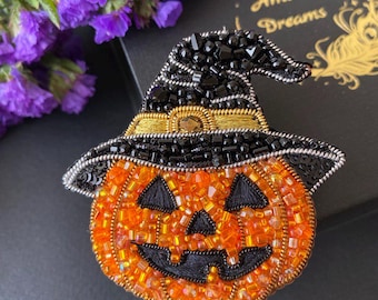 Może przedstawiać: Ręcznie robiona broszka na Halloween przedstawiająca pomarańczową dynię z uśmiechniętą twarzą, ozdobioną pomarańczowymi i czarnymi koralikami. Dynia nosi czarny kapelusz czarownicy ze złotymi wykończeniami.  Świąteczny dodatek na sezon jesienny.