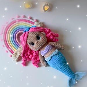 Pu&ograve; includere: Bambola sirena all'uncinetto fatta a mano con capelli rosa, coda blu e top a conchiglia. La bambola &egrave; sdraiata accanto a un'applique a forma di arcobaleno. Lo sfondo &egrave; bianco con luci decorative.