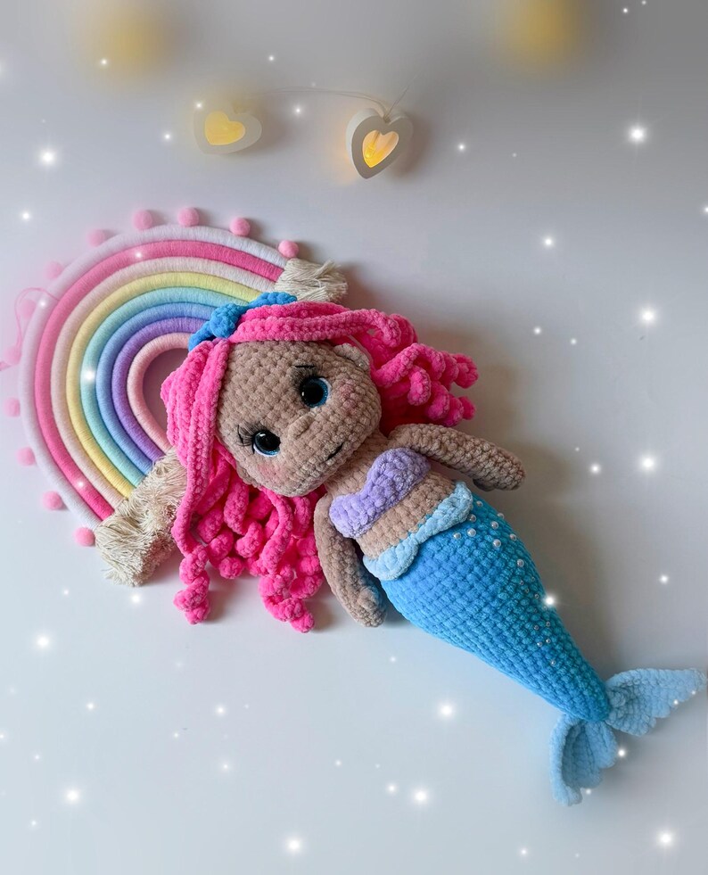 Pu&ograve; includere: Bambola sirena all'uncinetto fatta a mano con capelli rosa, coda blu e top a conchiglia. La bambola &egrave; sdraiata accanto a un'applique a forma di arcobaleno. Lo sfondo &egrave; bianco con luci decorative.