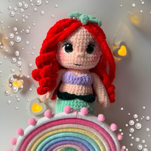Pu&ograve; includere: Una bambola sirena all'uncinetto fatta a mano con capelli rossi vibranti, un top viola e una coda verde. La bambola &egrave; appoggiata su un arcobaleno colorato con pompon rosa. Lo sfondo presenta luci decorative e bolle simulate.