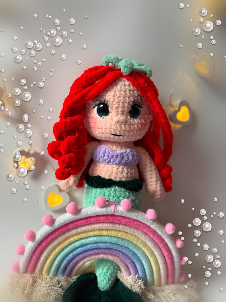 Pu&ograve; includere: Una bambola sirena all'uncinetto fatta a mano con capelli rossi vibranti, un top viola e una coda verde. La bambola &egrave; appoggiata su un arcobaleno colorato con pompon rosa. Lo sfondo presenta luci decorative e bolle simulate.