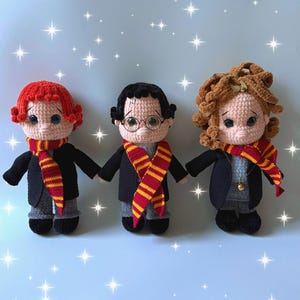 May include: Three handmade crochet dolls, each with a unique hairstyle and wearing a black jacket, gray pants, and a striped scarf in red and gold. The dolls are holding hands, and the background is a light blue with white star accents.