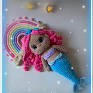 Pu&ograve; includere: Bambola sirena all'uncinetto fatta a mano con capelli rosa, coda blu e top a conchiglia. La bambola &egrave; sdraiata accanto a un arazzo arcobaleno. Lo sfondo &egrave; bianco con luci decorative.