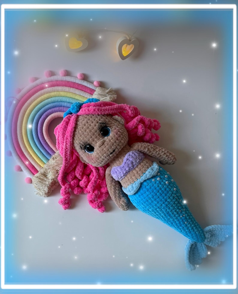 Pu&ograve; includere: Bambola sirena all'uncinetto fatta a mano con capelli rosa, coda blu e top a conchiglia. La bambola &egrave; sdraiata accanto a un arazzo arcobaleno. Lo sfondo &egrave; bianco con luci decorative.