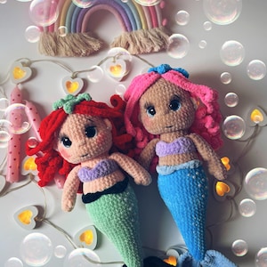 Pu&ograve; includere: Due bambole sirena all'uncinetto fatte a mano con capelli rossi e rosa, che indossano top viola e code verdi e blu. Un arazzo arcobaleno e luci a stringa con lampadine a forma di cuore sono sullo sfondo. Bolle fluttuano intorno alle bambole.