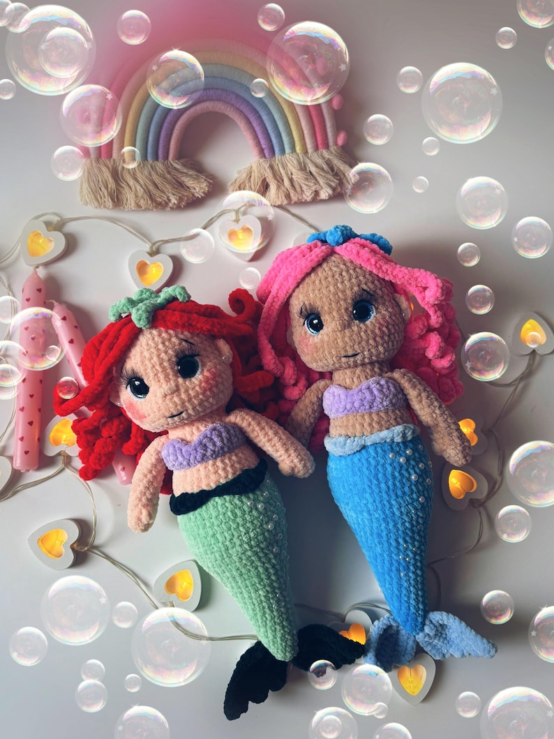 Pu&ograve; includere: Due bambole sirena all'uncinetto fatte a mano con capelli rossi e rosa, che indossano top viola e code verdi e blu. Un arazzo arcobaleno e luci a stringa con lampadine a forma di cuore sono sullo sfondo. Bolle fluttuano intorno alle bambole.