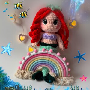Pu&ograve; includere: Bambola sirena all'uncinetto fatta a mano con capelli rossi, top lavanda e coda verde, posizionata sopra un arcobaleno. La scena include stelle marine, pesci e una medusa decorativi.