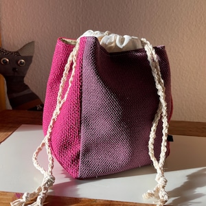 Könnte beinhalten: Ein zweifarbiges Kordelzugbeutel in Fuchsia- und Pflaumentönen. Die Tasche hat ein strukturiertes Gewebe und einen cremefarbenen Kordelverschluss. Ein Stofftier in Form einer Katze ist im Hintergrund zu sehen.