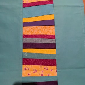 Könnte beinhalten: Ein türkisfarbenes Stoffpanel mit einem vertikalen Streifen aus buntem Stoff, der darauf genäht ist. Der Streifen besteht aus gelbem, violettem, türkisfarbenem, burgunderfarbenem und orangefarbenem Stoff mit Blumenmustern.