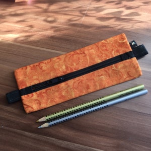 Könnte beinhalten: Orangefarbene Stiftetui mit schwarzem Reißverschluss und schwarzem Trageriemen. Das Etui ist aus Stoff mit einem Blumenmuster. Zwei Stifte liegen auf dem Etui.