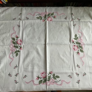 Könnte beinhalten: Weiße Tischdecke mit gestickten Rosen und Bändern in Rosa und Grün. Die Tischdecke ist quadratisch und mit einem Blumenmuster verziert.