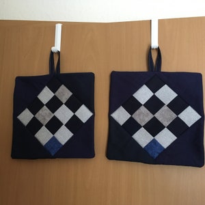 Könnte beinhalten: Zwei dunkelblaue Topflappen mit einem karierten Muster in Grau und Weiß. Die Topflappen hängen an weißen Haken an einer Holzoberfläche.