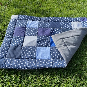 Könnte beinhalten: Ein Patchwork-Haustierbett mit blau-weißem Pfotenmuster, verschiedenen blauen Quadraten und einer grauen Unterseite. Das Bett ist rechteckig und liegt auf grünem Gras.