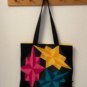 Könnte beinhalten: Schwarze Tragetasche mit vier farbenfrohen Sternenmustern in Pink, Gelb und Türkis. Die Tasche hat schwarze Träger und eine gelbe Innenverkleidung. Die Tasche hängt an einem Holzregal.