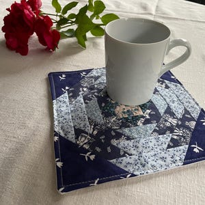 Könnte beinhalten: Eine weiße Keramiktasse steht auf einem quadratischen, gesteppten Untersetzer mit einem blau-weißen Patchwork-Design. Der Untersetzer hat einen dunkelblauen Rand mit weißen floralen Akzenten. Rote Rosen sind im Hintergrund.