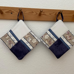 Könnte beinhalten: Zwei quadratische, gesteppte Topflappen mit einem Patchwork-Design aus weißen, dunkelblauen und beigen Stoffbahnen. Die beigen Abschnitte zeigen ein Muster aus verschiedenen Muscheln. Jeder Topflappen hat eine dunkelblaue Stoffschlaufe.