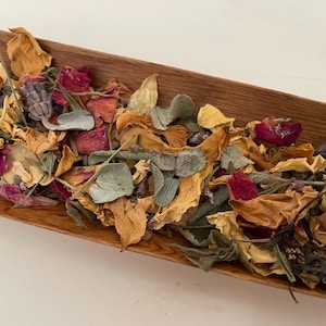 Könnte beinhalten: Eine Holzschale gefüllt mit einer bunten Auswahl an getrockneten Blumen und Kräutern. Die Anordnung umfasst orange, gelbe und rote Blütenblätter sowie grüne Blätter und Lavendelzweige. Die Schale ist aus hellbraunem Holz gefertigt.